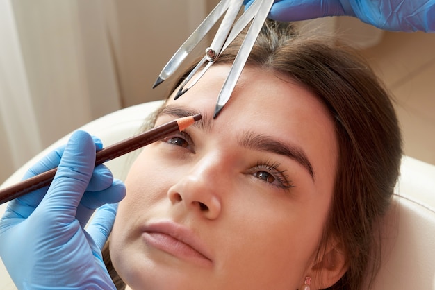 Mudando o formato das sobrancelhas. Estilista medindo as sobrancelhas com a régua. Fluxo de trabalho de micropigmentação em um salão de beleza. Mulher com as sobrancelhas tingidas com maquiagem semi-permanente.