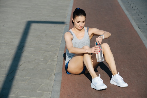 Müde athletische Frau, die draußen auf der Strecke sitzt und eine Flasche Wasser öffnet. An einem sonnigen Tag unter strahlend blauem Himmel. Sie trägt ein weißes ärmelloses Hemd und Mini-Shorts.