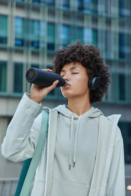 Müde, athletische Frau in Jacke und Hoodie erfrischt sich mit Energy-Drink nach aktivem Joggen, hört Musik über Stereo-Kopfhörer-Posen vor verschwommenem Gebäudehintergrund und hat tägliche Übungsroutine