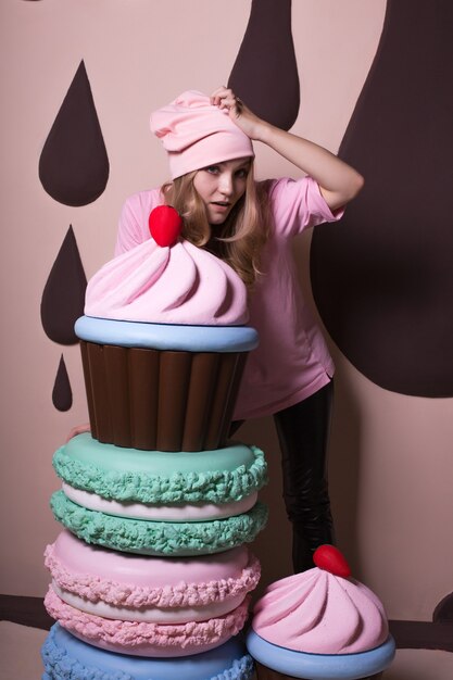 Müde blonde Frau mit rosa Mütze und T-Shirt, die Spaß mit großen Makronen und Cupcakes im Studio hat