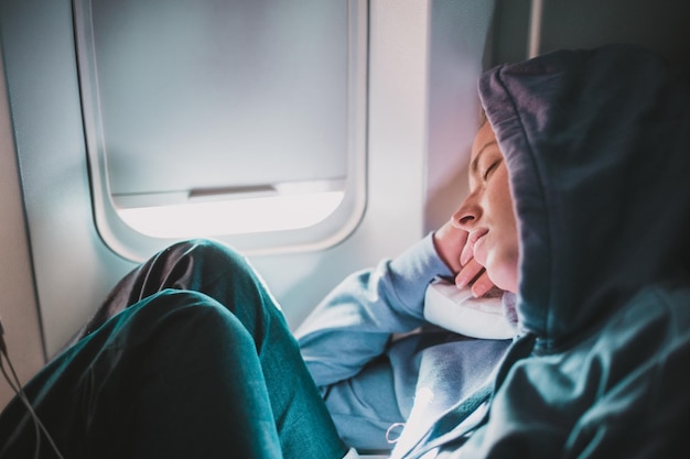 Müde blonde, legere kaukasische Frau mit sportlichem Kapuzenpullover schläft auf dem Sitz, während sie mit dem Flugzeug reist