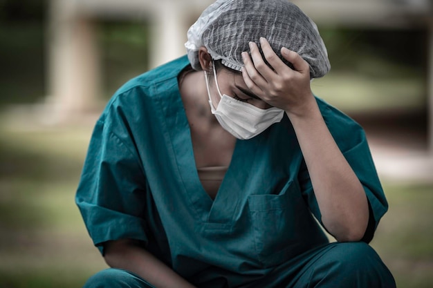 Müde depressive asiatische Peeling-Krankenschwester trägt Gesichtsmaske blaue Uniform sitzt auf dem Krankenhausboden Junge Ärztin gestresst von harter Arbeit