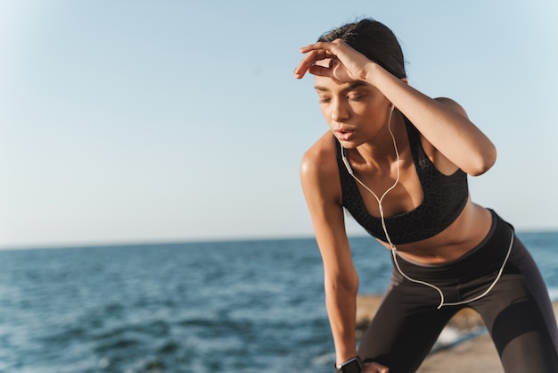 müde ernsthafte schöne junge starke sportfrau draußen am strand morgens ruhen sie sich aus und hören sie musik mit kopfhörern.