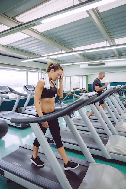 Müde Frau trocknet mit Handtuch beim Training auf einem Laufband im Fitnesscenter