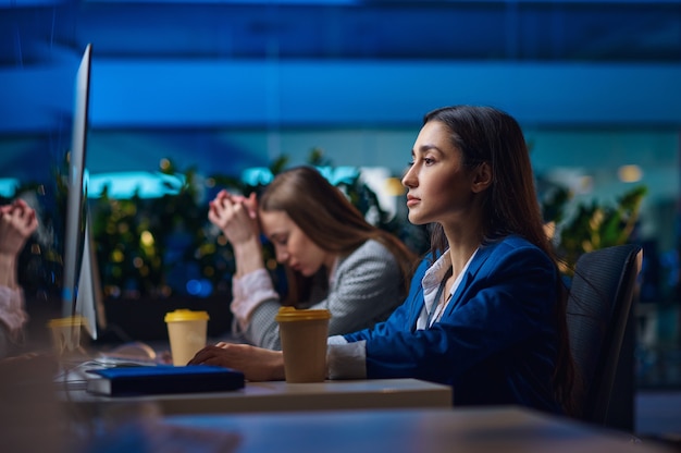 Müde Frauen sitzen am Tisch im Nachtbüro. Schläfrige Geschäftsfrauen, dunkles Geschäftszentrum, moderner Arbeitsplatz