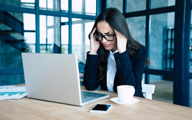 Müde Geschäftsfrau in vollem Anzug mit Kopfschmerzen beim Arbeiten an ihrem Laptop im modernen Büro hält ihren Kopf