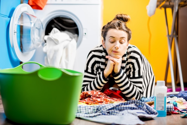 Müde Hausfrau sitzt auf dem Boden neben der Waschmaschine mit bunten Klamotten zu Hause