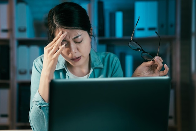 Müde junge asiatische japanische Freiberuflerin, die an Laptop-Computer mit Deadline-Projekt im Home Office arbeitet. Mädchen in Freizeitkleidung, die Überstunden am Notebook-PC macht, fühlt sich gestresst und schmerzt in den Augen.