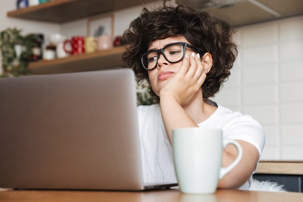 müde junge Frau, die drinnen an der Küche mit Laptop-Computer sitzt