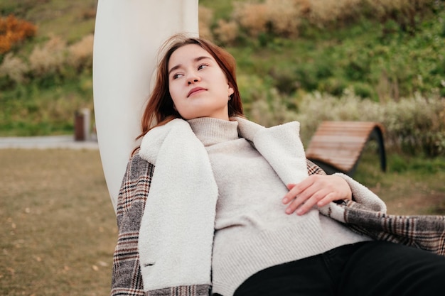 Müde junge Frau im Mantel, die sich im Herbstpark auf eine weiße moderne Schaukel legt Müdigkeit nach der Arbeit Entspannendes Chillen im Freien