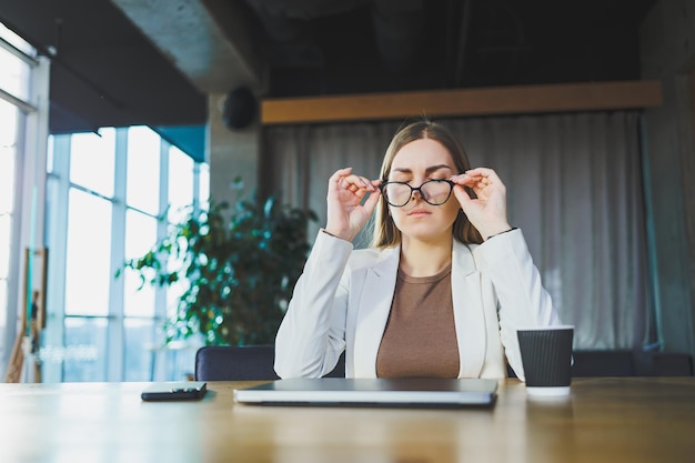 Müde junge Geschäftsfrau, die im Büro an einem Holztisch hinter einem Laptop sitzt Ein junger Manager mit Brille ist müde von der Arbeit