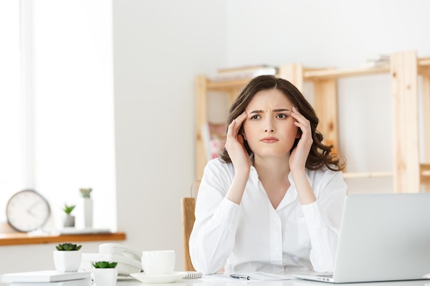 Müde junge Geschäftsfrau, die unter langer Zeit leidet, die am Computertisch im Büro sitzt