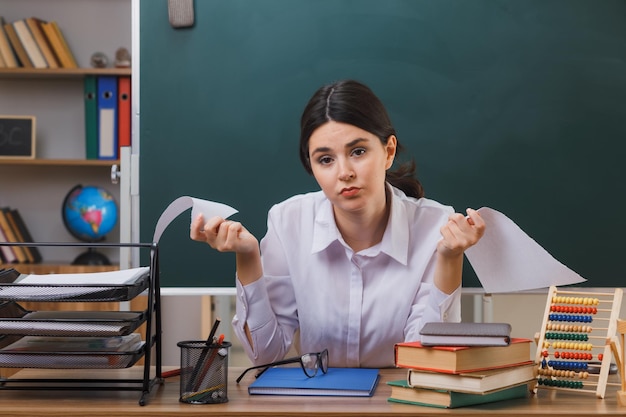 müde junge reißpapierlehrerin, die am schreibtisch mit schulwerkzeugen im klassenzimmer sitzt