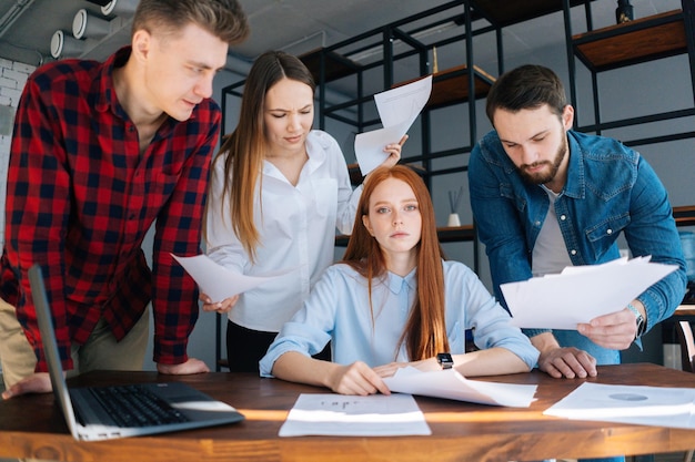 Müde junge weibliche Teamleiterin unter Stress im Büro am Schreibtisch sitzend mit gemischtrassigen Kollegen, die Papierdokumente schütteln, die neben ihr stehen und in die Kamera schauen