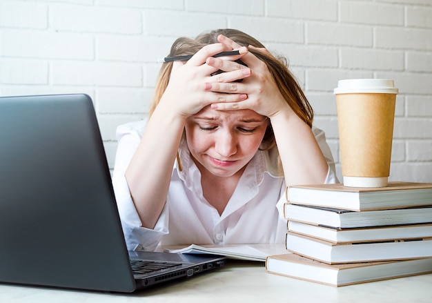 Müde Mädchen hält seinen Kopf mit den Händen und sitzt am Schreibtisch zu Hause und lernt am Laptop mit Büchern herum.