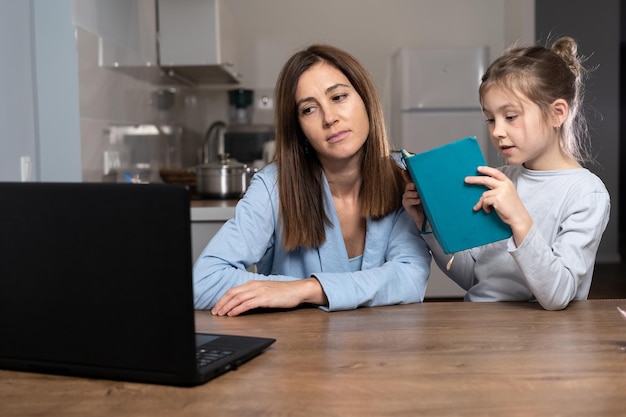 Müde Mutter mit traurigem Blick hilft dem Kind bei den Hausaufgaben sitzt am Tisch neben dem Laptop Tochter liest fröhlich Text aus einem Notizbuch