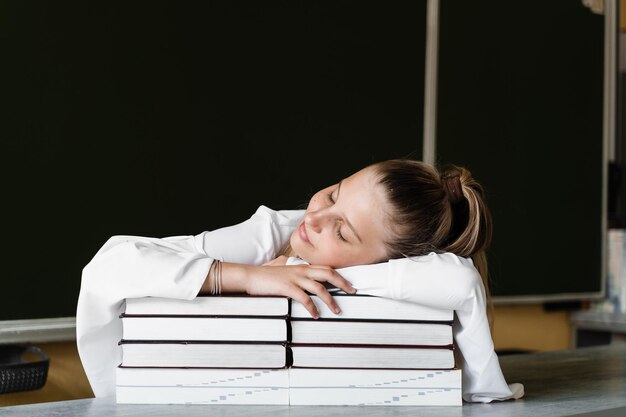 Müde Schulmädchen liegt und schläft auf Büchern an der Tafel in der Schule Schulbildung Das Mädchen ist es leid, Hausaufgaben zu unterrichten