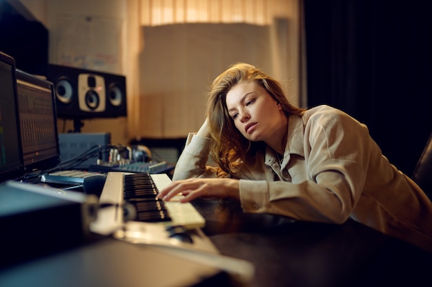 Müde Toningenieurin in Kopfhörern, Aufnahmestudio-Interieur im Hintergrund. Synthesizer und Audiomixer, Musikerarbeitsplatz, harter kreativer Prozess