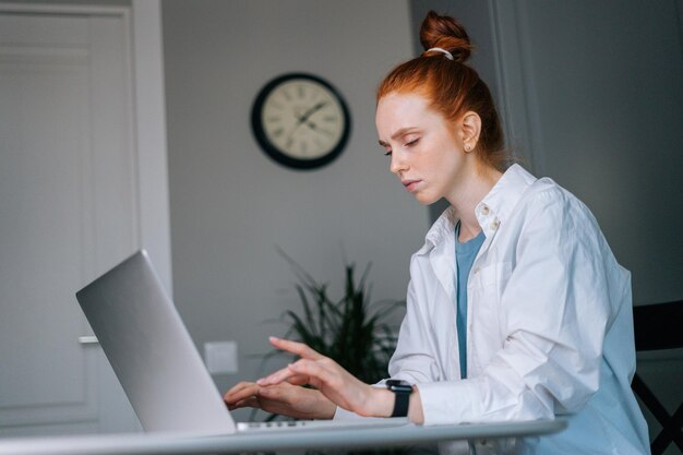 Müde, überarbeitete, rothaarige junge Frau, die am Tisch im Wohnzimmer am Laptop-Computer tippt. Konzept der Freizeitbeschäftigung rothaarige Frau zu Hause während der Selbstisolation.