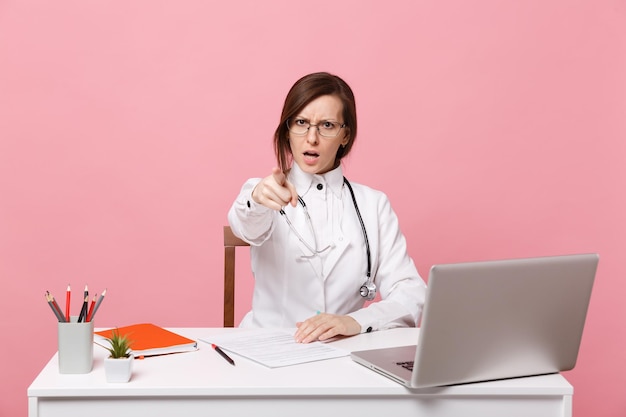 Müde verrückte Ärztin sitzt an der Schreibtischarbeit am Computer mit medizinischem Dokument im Krankenhaus einzeln auf pastellrosa Wandhintergrund. Frau im medizinischen Kittelbrillenstethoskop. Konzept der Gesundheitsmedizin.