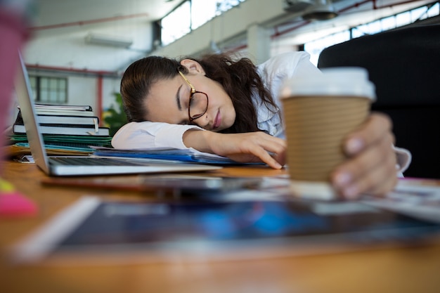 Müde weibliche Grafikdesignerin, die Einwegbecher beim Schlafen auf Schreibtisch hält