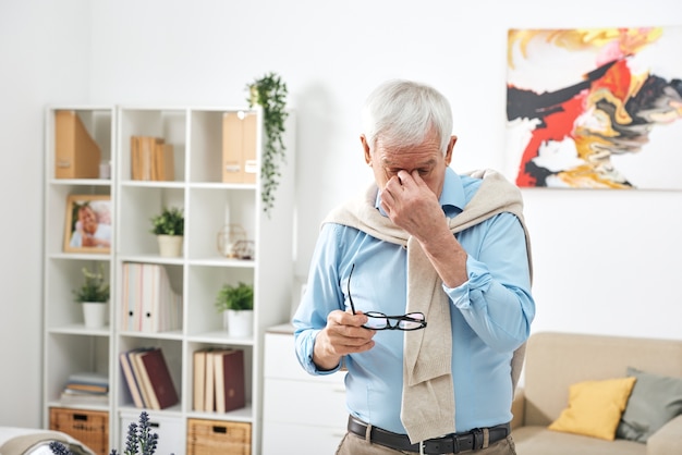 Müder älterer Mann im blauen Hemd, das Brillen hält und Nasenrücken reibt, während Augenmüdigkeit zu Hause fühlt