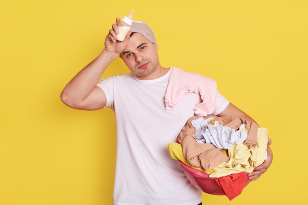 Müder, erschöpfter Ehemann, der mit Babyartikeln isoliert über gelber Wand steht, hält Vater die Flasche in den Händen und hält die Hand auf der Stirn.