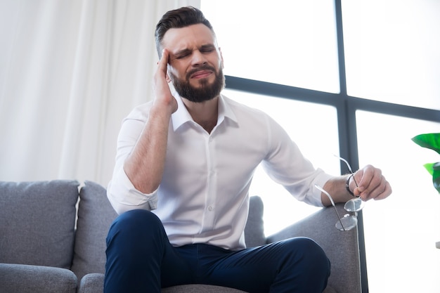 Müder Geschäftsmann mit Stress hat Kopfschmerzen und sitzt mit Schmerzen im Gesicht auf der Couch