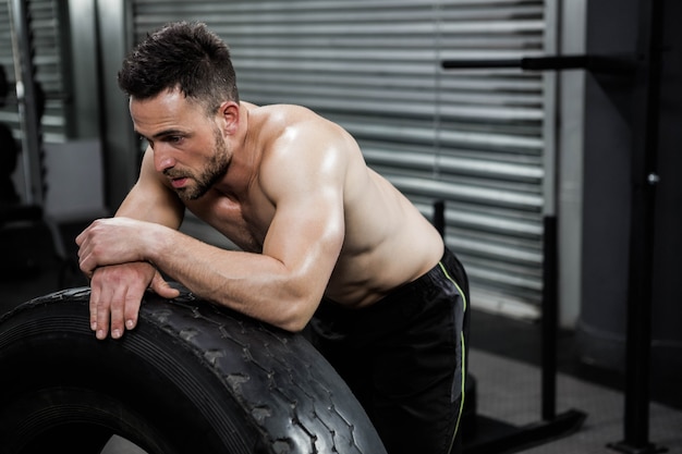 Müder hemdloser Mann, der am schweren Reifen an der crossfit Turnhalle sich lehnt