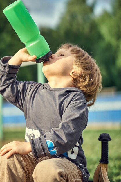 Müder Junge mit Holzlaufrad trinkt Wasser aus Plastikflasche, während er auf grünem Gras sitzt