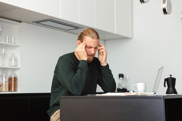 Müder junger bärtiger Mann mit Kopfschmerzen, die an der Küche mit Laptop-Computer sitzen.