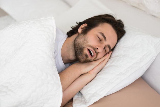 Müder junger Kaukasier mit offenem Mund, der in der Freizeit auf einem weißen, weichen Bett im Schlafzimmer schläft, genießt die Ruhe