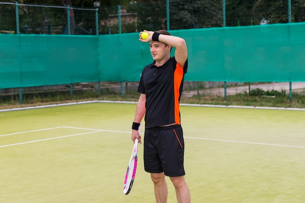 Müder junger männlicher Tennisspieler, der eine Sportbekleidung trägt, die Tennisball und Schläger nach dem Spiel auf einem Platz im Freien im Sommer hält