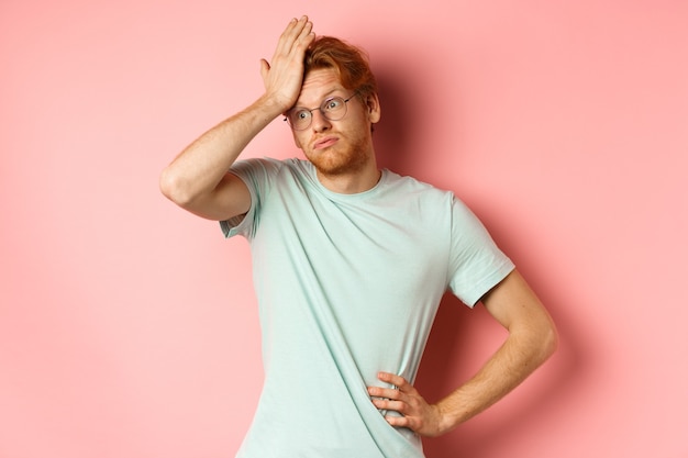 Müder junger Mann mit roten Haaren, der eine Brille trägt und genervt und angespannt aussieht und eine Facepalm-Geste macht und...