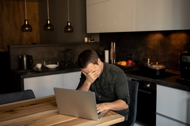 Müder Mann hält seinen Kopf, während er drinnen an einem Laptop arbeitet. Online-Mitarbeiter, Freiberufler am Heimarbeitsplatz - Küchenzimmer