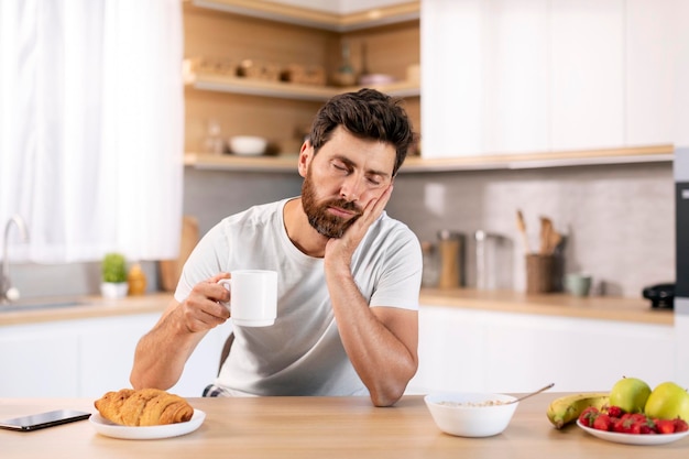 Müder, trauriger, schläfriger, gutaussehender tausendjähriger Kaukasier mit Bart in Hauskleidung mit einer Tasse Kaffee schläft am Tisch