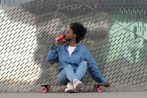 Müdes afroamerikanisches Skateboardermädchen, das sich nach dem Longboarden erfrischt, sitzt auf einem Skate-Drink-Soda-Getränk