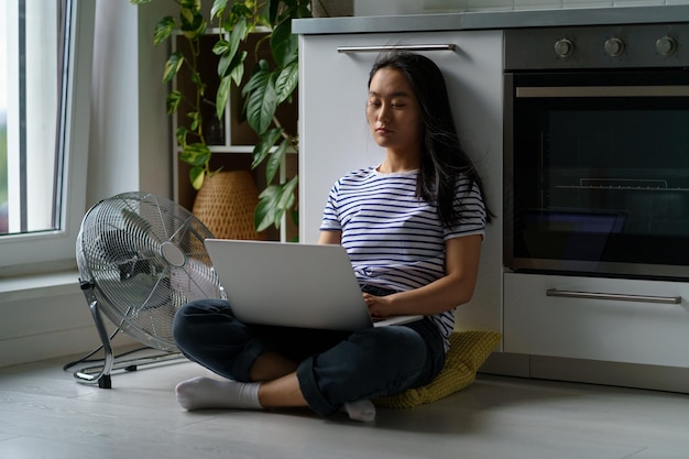 Müdes asiatisches Mädchen sitzt mit Laptop auf dem Boden und hat einen Ventilator, leidet unter der Hitze zu Hause und versucht sich zu konzentrieren