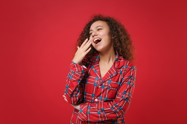 Müdes junges afrikanisch-amerikanisches Mädchen in Pyjama-Homewear posiert, während es sich zu Hause isoliert auf rotem Hintergrund ausruht. Entspannen Sie sich bei guter Laune Lifestyle-Konzept. Mock-up-Kopienbereich. Gähnen, den Mund mit der Hand bedeckend.