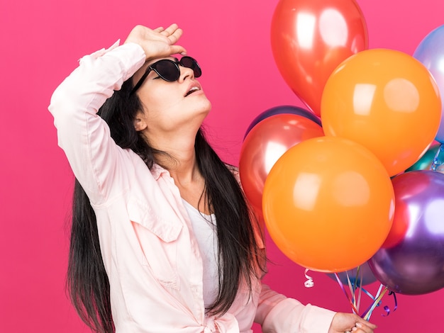 Müdes junges schönes mädchen mit brille, das ballons hält und hand auf die stirn legt