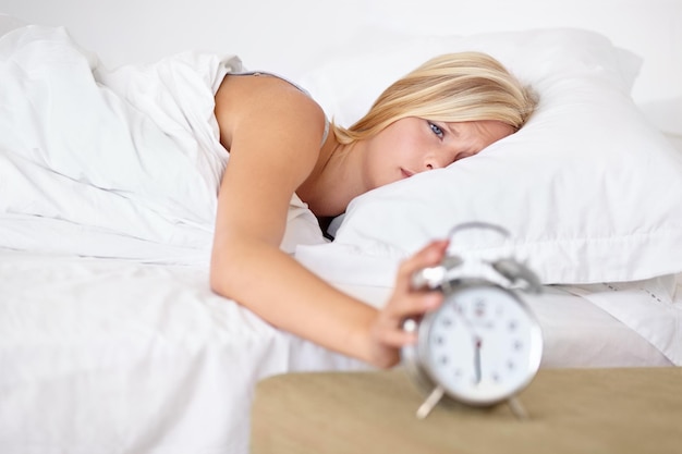 Müdige Frau und aufwachen mit Alarm am Morgen und schlafen auf dem Bett zu Hause mit Stress oder Müdigkeit Drücken Sie Snooze und Mädchen frustriert mit Uhr und Zeit, um den Tag in Schlafzimmer und Wohnung zu beginnen