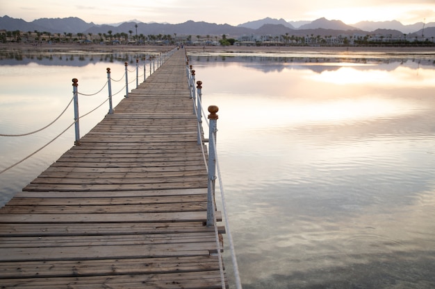 Muelle de madera largo entre el mar con montañas en el horizonte al atardecer.