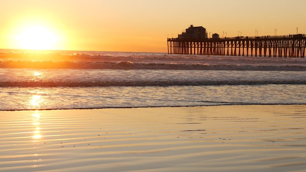 Muelle de madera sobre pilotes, silueta al atardecer, California, Estados Unidos, Oceanside. Olas del mar soleado al atardecer.