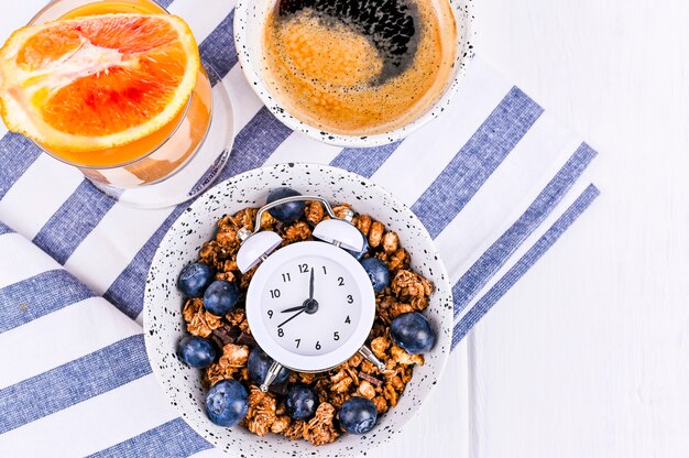 Muesli con arándanos en una taza, jugo de naranja y aromático café matutino. Desayuno sobre un fondo blanco de madera y un reloj despertador. Endecha plana. Copia espacio