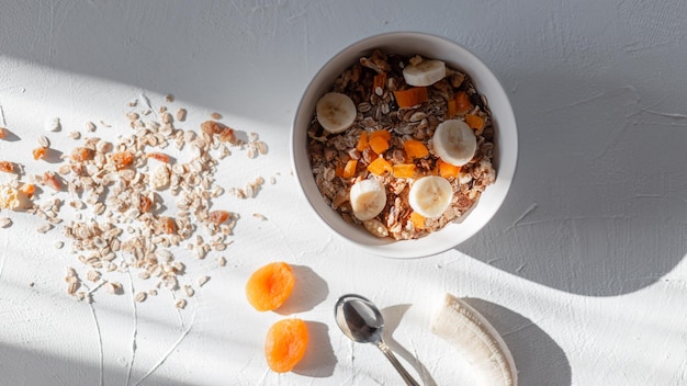Muesli de desayuno de alimentos crudos saludables con avena, albaricoques secos, plátano y frutas secas en una mesa blanca al amanecer por la mañana, vista superior.