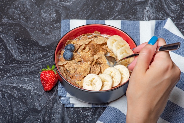 Müsli. Frühstück, gesunde Ernährung und Ernährung. Müsli mit Milch und Früchten in einem Teller auf einer schwarzen Marmorplatte. Frauenhand mit einem Löffel