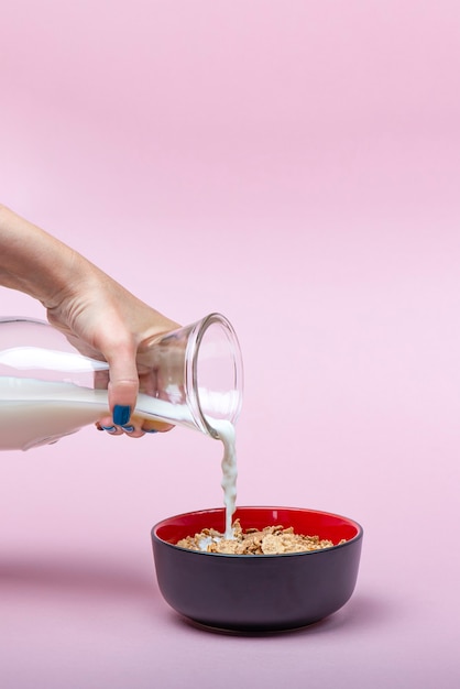 Müsli kochen. Eine Frau gießt Milch aus einem Krug in eine Untertasse mit Müsliflocken. Rosa Hintergrund mit Platz zum Einfügen von Text.