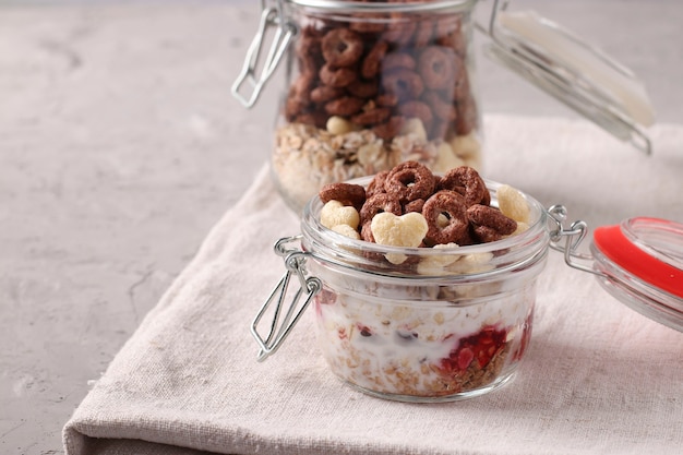 Muesli de miel crujiente de granola con frutos rojos, requesón y yogur natural, ubicado en dos frascos de vidrio sobre un fondo gris, formato horizontal, espacio para texto