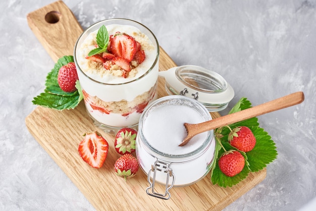 Müsli oder Joghurt mit Erdbeere im Glas, frischen Beeren und Glas mit Zucker auf einem Schneidebrett, Draufsicht