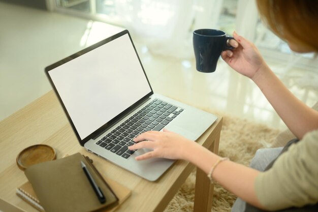 Muestra cortada de una mujer joven sosteniendo una taza de café y usando una computadora portátil en la sala de estar